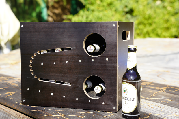 Holzkasten mit zwei runden Ausschnitten für Flaschen, daneben eine Flasche 'Krombacher Pils'. Der Kasten hat eine geschwungene Linie und Löcher auf der Vorderseite.