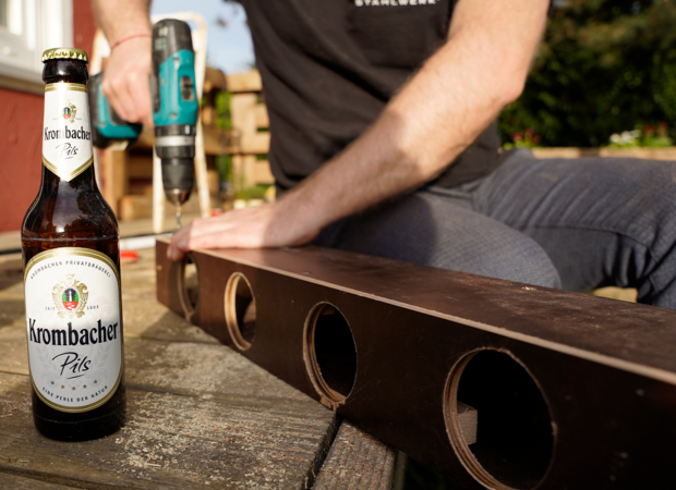 Fünf Getränkehalter werden gebaut und Holzplatten zusammengeschraubt. Eine Krombacher Pilsflasche steht daneben.