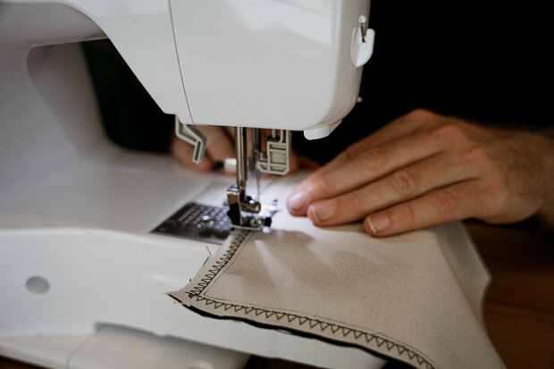 Eine Person näht mit einer Nähmaschine an einem hellen Stoffstück, das mit einer Zickzacklinie verziert ist. Die Hände der Person sind sichtbar, während sie den Stoff führt.