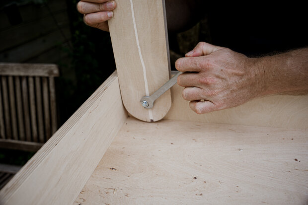 Eine Hand hält einen Holzpfosten, während die andere Hand einen Schraubenschlüssel an einer Metallmutter anwendet. Der Tisch ist aus hellem Holz gefertigt und hat eine klare Maserung.