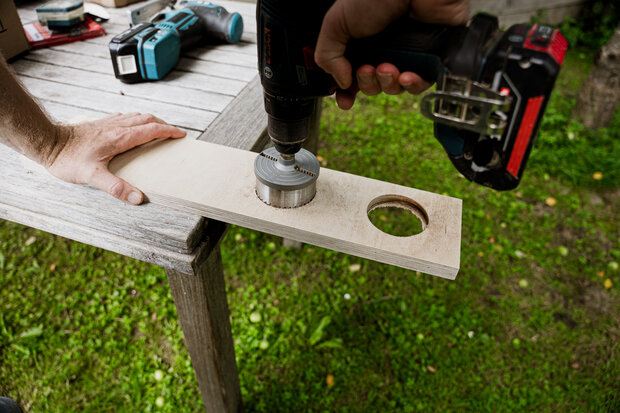 Eine Hand hält einen Akku-Bohrer mit einem Lochsäge-Aufsatz, der auf ein Holzbrett gerichtet ist. Das Brett liegt auf einem Tisch im Freien, mit einer kreisförmigen Aussparung und einem weiteren Werkzeug im Hintergrund.