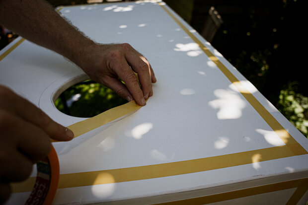 Eine Hand zieht gelbes Klebeband über die Kante einer weißen Oberfläche mit einem runden Ausschnitt. Die Szene ist von Schatten durch Blätter umgeben.