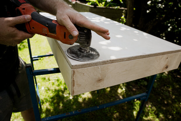 Eine Person mit einem schwarzen T-Shirt benutzt einen elektrischen Schleifer, um die Kante einer Holzplatte zu glätten. Die Holzplatte liegt auf einem blauen Gestell im Freien.