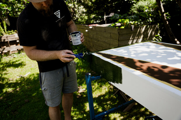 Ein Mann mit Bart trägt ein schwarzes T-Shirt und graue Shorts, während er mit einem Pinsel grüne Farbe auf die Kante eines weißen Tisches aufträgt. Im Hintergrund sind Pflanzen und ein Holzgestell sichtbar.
