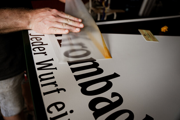 Eine Hand platziert eine transparente Folie auf einem weißen Schild mit dem Text 'Jeder Wurf ein Treffer'. Der Fokus liegt auf der präzisen Positionierung der Folie.