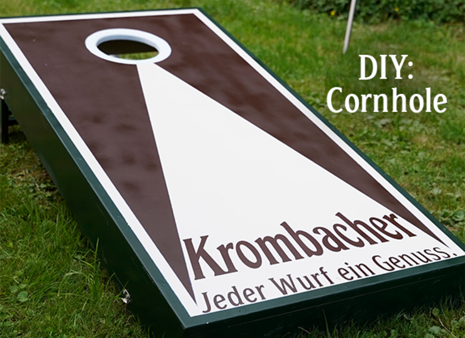 Ein Cornhole-Spielbrett mit einem großen, runden Loch in der oberen Mitte. Auf dem Brett steht 'Krombacher' und der Slogan 'Jeder Wurf ein Genuss.'
