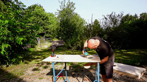 Ein Mann mit Glatze arbeitet an einem Holztisch im Freien und benutzt eine Bohrmaschine. Im Hintergrund sind Bäume und ein Zaun sichtbar.