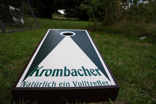 Ein Krombacher Cornhole-Spielbrett liegt auf dem Gras. Es hat eine grüne und weiße Farbgestaltung mit dem Text 'Krombacher' und 'Natürlich ein Volltreffer'.