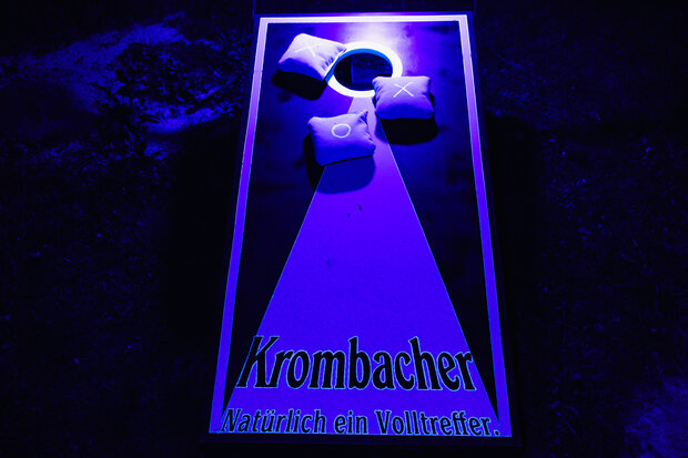 Ein Krombacher-Werbeschild mit einem weißen Dreieck, das auf eine Fläche mit zwei weißen Kissen zeigt. Der Text lautet: 'Krombacher Natürlich ein Volltreffer.'