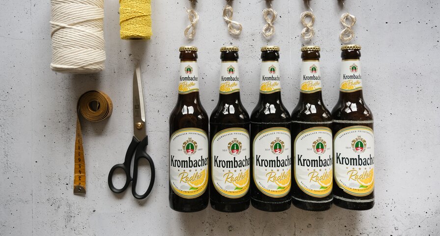 Fünf Flaschen Krombacher Radler stehen in einer Reihe. Daneben liegen eine Schere, ein Maßband, zwei Rollen Schnur in Weiß und Gelb sowie vier Korken mit Clips.