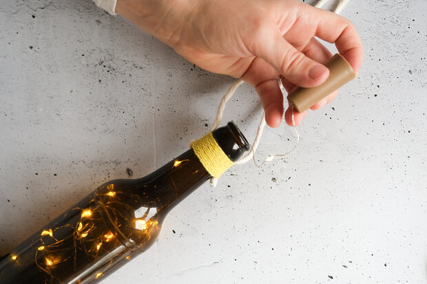 Eine Hand hält einen Korken, der auf eine braune Glasflasche mit Lichtern im Inneren passt. Die Flasche hat einen gelben Faden um den Hals gewickelt.