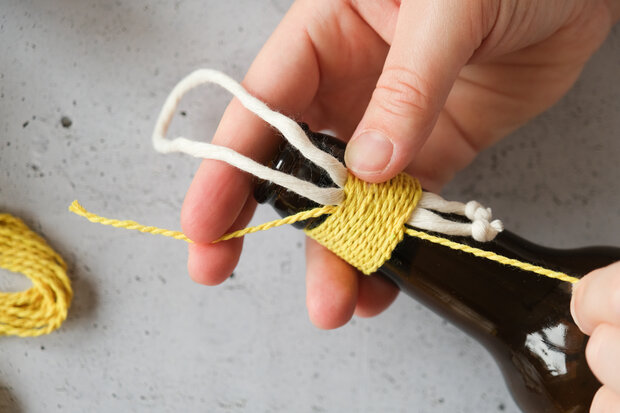 Eine Hand wickelt gelbes Garn um den Hals einer braunen Flasche. Neben der Flasche liegen weitere gelbe und weiße Schnüre.