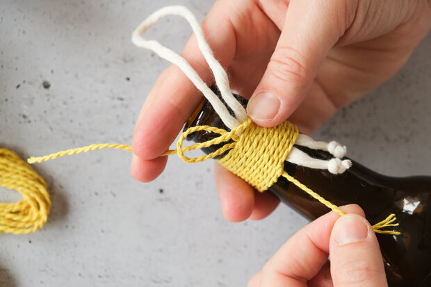 Eine Person wickelt gelbes Garn um den Hals einer braunen Flasche. Zwei weiße Fäden sind sichtbar, die aus dem Garn herausragen.
