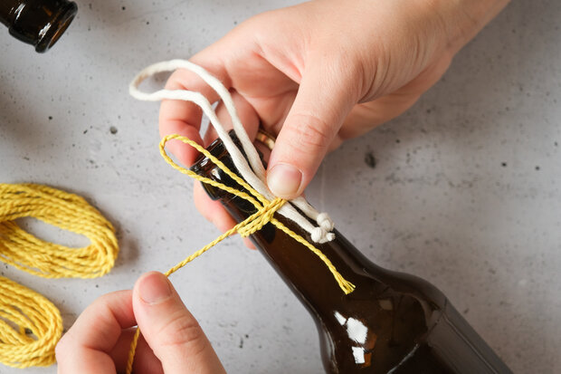 Eine Person bindet gelbe und weiße Schnüre um den Hals einer braunen Flasche. Im Hintergrund sind weitere Flaschen und Schnüre sichtbar.