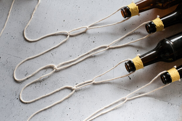 Fünf braune Flaschen mit gelben Kordeln am Hals liegen auf einer grauen Oberfläche. Zwischen den Flaschen verlaufen mehrere weiße Schnüre.