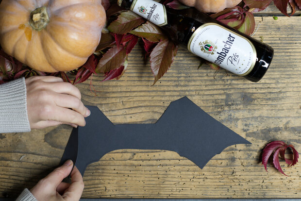 Schwarzes eckig ausgeschnittenes Papier mit Kürbis und einer bierflasche auf einem Holztisch.