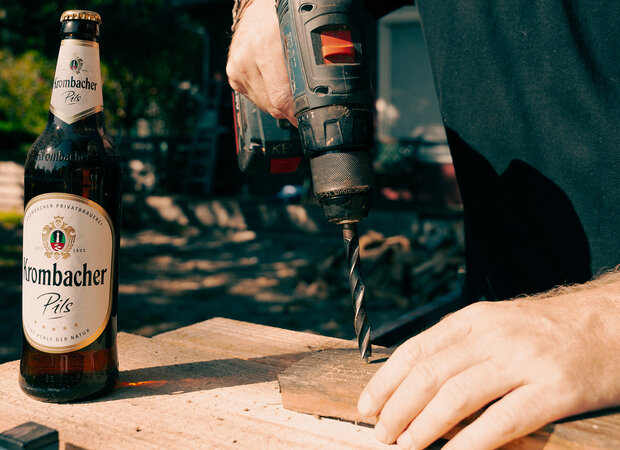 In die Daube werden Löcher gebohrt. Daneben steht eine Krombacher Pils Flasche.