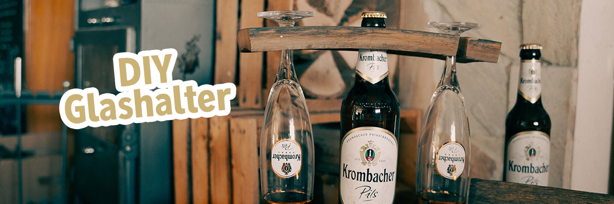 Eine Pilsflasche trägt am Flaschenhals ein Holzstück mit zwei Krombacher Pils Gläsern und Text: DIY Glashalter.