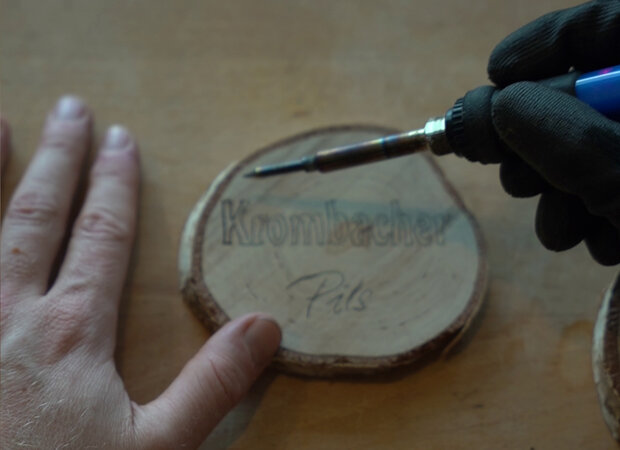 Mit einem Lötkolben wird das Krombacher Pils Branding in die Holzscheibe reingefräst.
