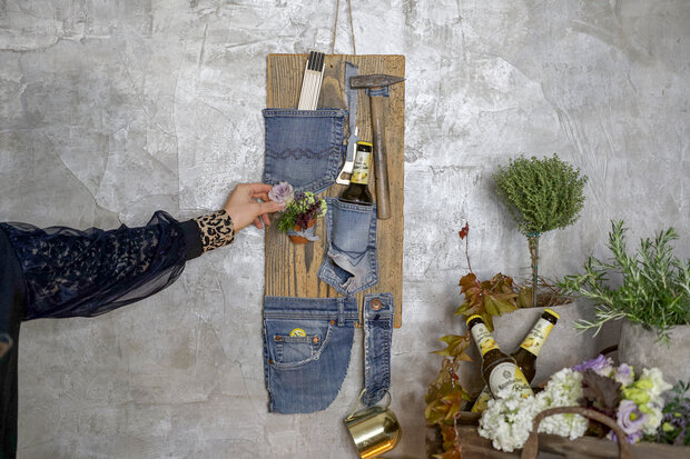 Eine Person mit einem schwarzen Oberteil und einem gemusterten Ärmel greift nach einer Blume, die in einer Tasche aus Jeansstoff auf einem Holzbrett steckt. Das Brett ist mit verschiedenen Utensilien wie Besteck und einer Flasche Bier dekoriert. Im Vordergrund sind Pflanzen und Blumen in Töpfen zu sehen.