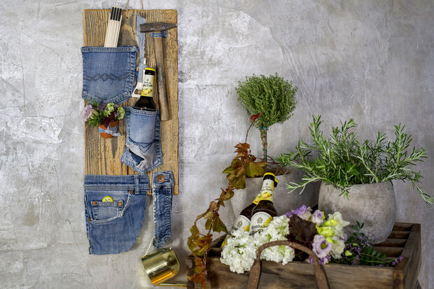 Holzbrett mit verschiedenen Taschen aus Jeans, gefüllt mit Besteck und Flaschen. Rechts eine Pflanze in einem grauen Topf und eine Holzkiste mit Blumen.