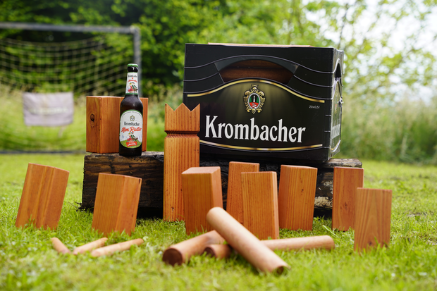 Krombacher AlmRadler Flasche mit einer Krombacher Pils Kiste und einem aufgestellten Wikingerschach-Kubb-Spiel.