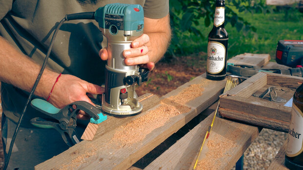 Eine Person mit einem blauen Handfräser bearbeitet ein Holzbrett. Auf dem Tisch stehen zwei Bierflaschen mit dem Etikett 'Erdinger'. Holzspäne liegen auf dem Tisch.
