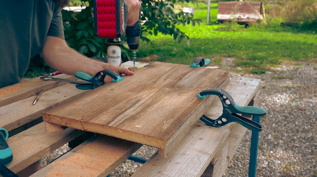 Eine Person mit einem Akkuschrauber bearbeitet ein Holzbrett, das auf einem Tisch fixiert ist. Zwei Klemmen halten das Brett an Ort und Stelle. Im Hintergrund sind Bäume und eine Bank sichtbar.