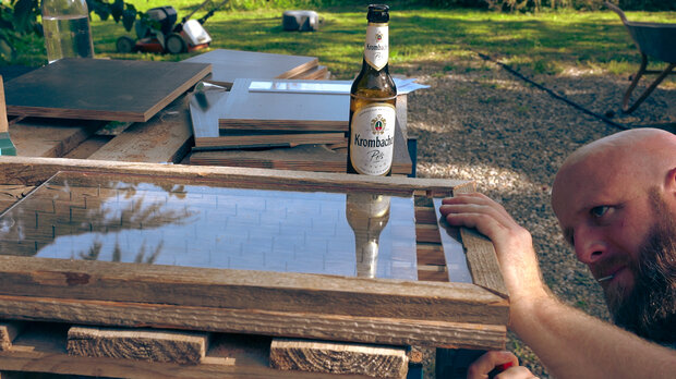 Ein Holzrahmen mit einem Spiegel wird auf einem Tisch gehalten. Im Hintergrund steht eine Flasche Kombucha. Der Tisch ist mit verschiedenen Holzplatten und Werkzeugen bedeckt.