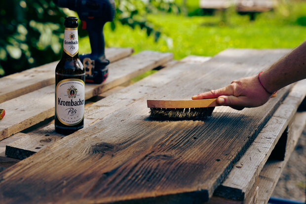 Ein Handwerker streicht mit einer Bürste über ein Holzbrett. Neben ihm steht eine Flasche Krombacher Pils. Im Hintergrund sind grüne Bäume und eine Holzplattform sichtbar.