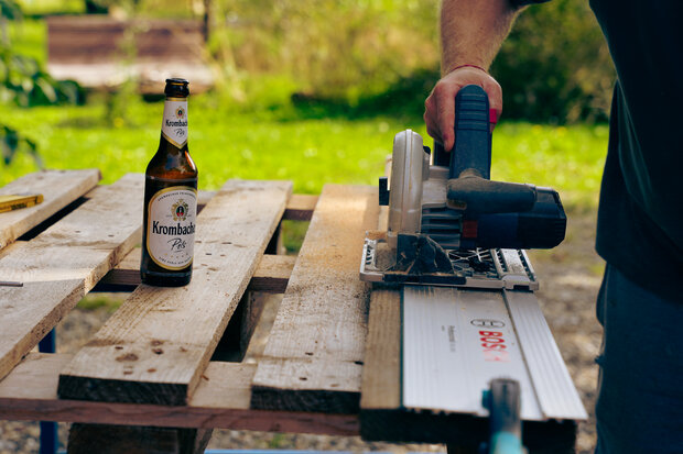 Eine Person schneidet ein Holzbrett mit einer elektrischen Kreissäge. Neben der Säge steht eine Flasche Krombacher Pils. Der Hintergrund zeigt eine grüne Wiese.