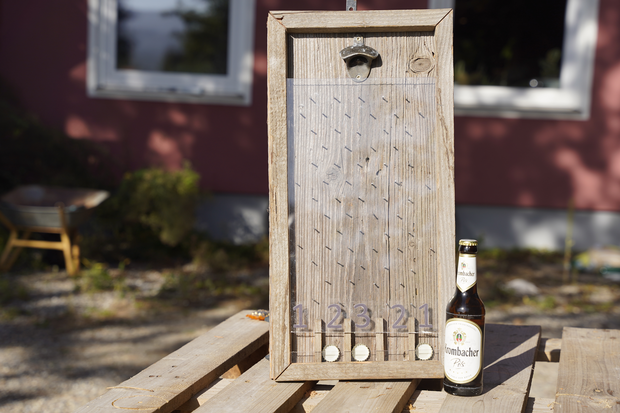 Ein hölzerner Flaschenöffner mit einer geriffelten Oberfläche und der Aufschrift '1.2.3.21' steht auf einem Tisch. Daneben steht eine Flasche 'Krombacher Pils', die aufrecht steht.