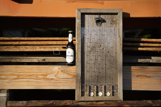 Ein hölzerner Flaschenöffner mit einem geflochtenen Muster, an einer Wand montiert. Neben dem Öffner steht eine Flasche Bier mit dem Etikett 'Erdinger'. Unter dem Öffner sind die Zahlen '1 2 3 4 5 6' sichtbar.