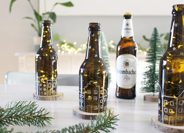 Drei braune Bierflaschen mit Lichtern in einem dekorativen Muster stehen auf Holzuntersetzern. Im Hintergrund steht eine Flasche 'Krombacher Pils'. Kleine grüne Tannenbäume sind ebenfalls sichtbar.