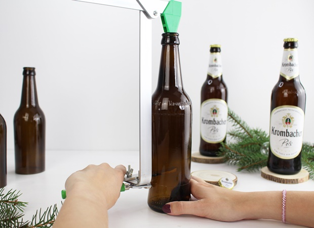 Eine Hand hält eine braune Glasflasche, während ein Flaschenöffner mit grünem Griff verwendet wird. Im Hintergrund stehen mehrere Krombacher Pils-Flaschen.