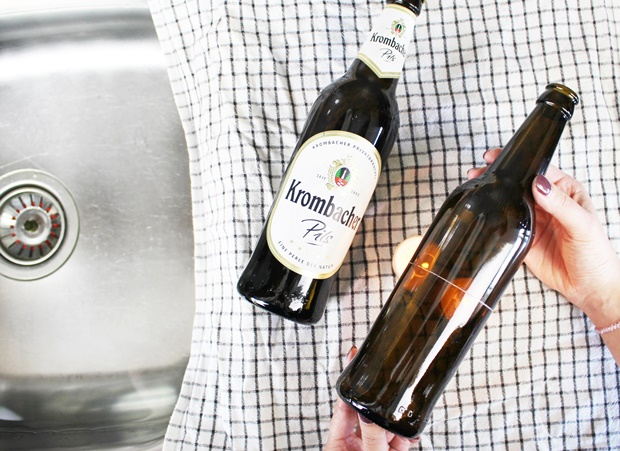 Zwei Flaschen Krombacher Pils stehen auf einem karierten Tuch. Eine Flasche wird von einer Hand gehalten, die andere liegt daneben. Im Hintergrund ist ein Spülbecken sichtbar.