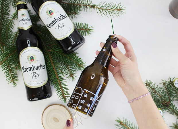 Eine Hand hält eine braune Flasche, die wie ein Haus mit Fenstern dekoriert ist. Im Hintergrund stehen zwei weitere Flaschen Krombacher Pils, umgeben von Tannenzweigen.