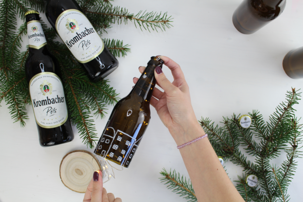 Eine Hand hält eine geöffnete Krombacher Pils Flasche, während zwei weitere Flaschen im Hintergrund stehen. Umgeben von Tannenzweigen und Holzscheiben.