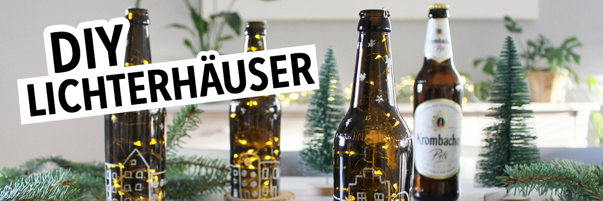 Drei braune Glasflaschen mit Lichtern, die wie kleine Häuser dekoriert sind, stehen auf einem Tisch. Im Hintergrund eine Flasche 'Krombacher Pils' und kleine grüne Tannenbäume. Oben steht der Text 'DIY LICHTERHÄUSER'.