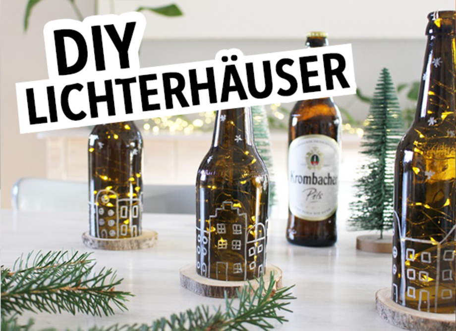 Drei braune Glasflaschen mit Lichtern, die wie kleine Häuser dekoriert sind, stehen auf einem Tisch. Im Hintergrund eine Flasche 'Krombacher Pils' und kleine grüne Tannenbäume. Oben steht der Text 'DIY LICHTERHÄUSER'.