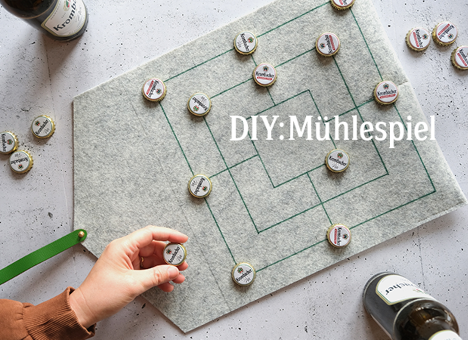 Eine Person mit braunen Haaren spielt Mühlespiel auf einem grauen Filzbrett mit grünen Linien und Flaschenverschlüssen als Spielfiguren. Sichtbar ist der Text 'DIY: Mühlespiel'.