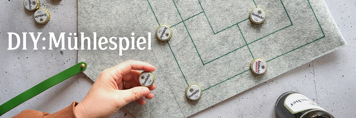 Eine Person mit braunen Haaren spielt Mühlespiel auf einem grauen Filzbrett mit grünen Linien und Flaschenverschlüssen als Spielfiguren. Sichtbar ist der Text 'DIY: Mühlespiel'.