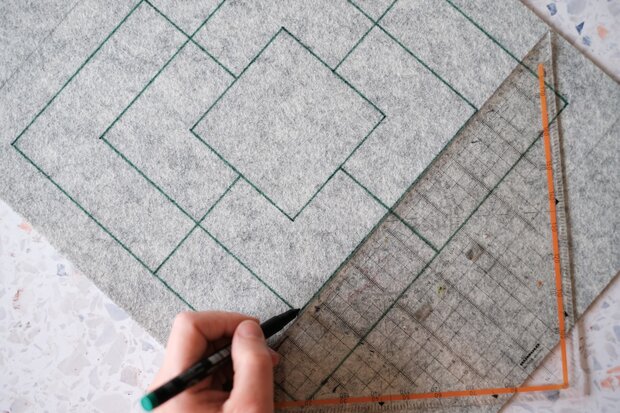 Ein Handwerker zeichnet mit einem schwarzen Marker auf grauem Filz ein geometrisches Muster mit mehreren Rauten. Ein transparentes Geodreieck liegt auf der Unterlage.