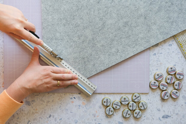 Eine Hand schneidet graues Filzmaterial mit einem Cutter entlang eines Lineals. Neben dem Filz liegen mehrere runde, bedruckte Knöpfe.