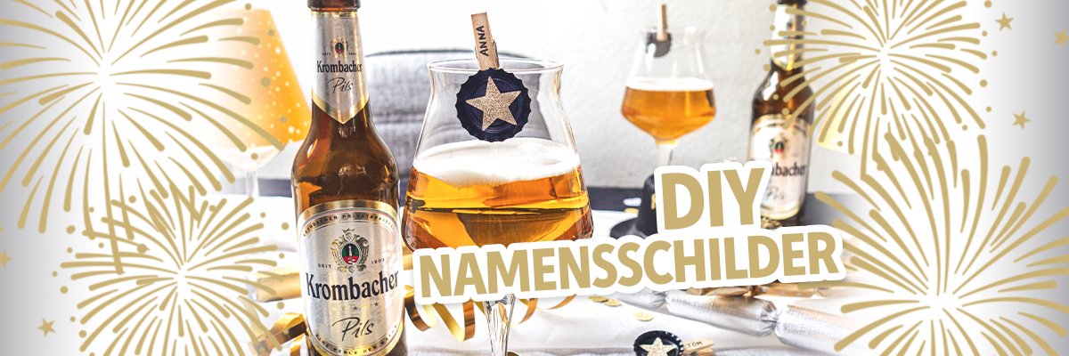 Ein Krombacher Pils Bier steht neben einem Glas mit einem goldenen Stern und einem Namensschild, das 'ANNA' zeigt. Auf dem Tisch sind Dekorationen für Silvester, darunter eine goldene Tischlampe und Konfetti.