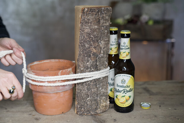 Ein Blumentopf mit zwei Holzstücken sind mit einer Kordel zusammen gebunden. Rechts davon stehen zwei Krombacher NaturRadler-Flaschen