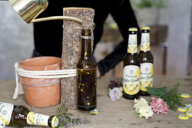 Ein Blumentopf, welcher mit einer Kordel und einem Holzstück zusammengebunden ist und einer braunen Flasche wird mit einer Gießkanne und Wasser gefüllt.