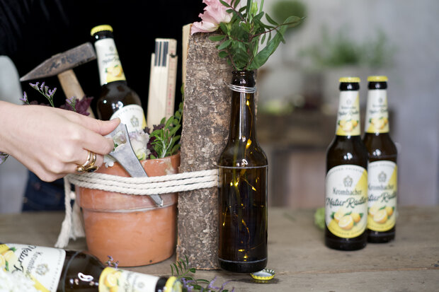 Blumentopf, der mit einer Kordel an ein Holzstück und eine Flasche gebunden ist, wird bestückt mit einem Hammer, einer Krombacher NaturRadler Flasche und einem Zollstock.