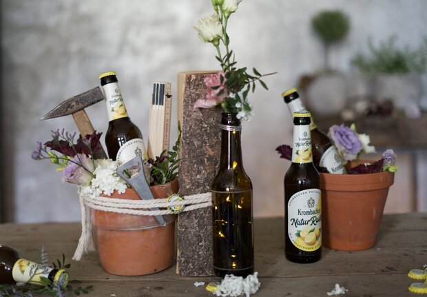 Zwei braune Bierflaschen von Krombacher mit dem Etikett 'Krombacher NaturRadler' stehen neben einem Holzblock und Tontöpfen. In den Töpfen sind Blumen und Werkzeuge wie eine kleine Schaufel und Bleistifte sichtbar.