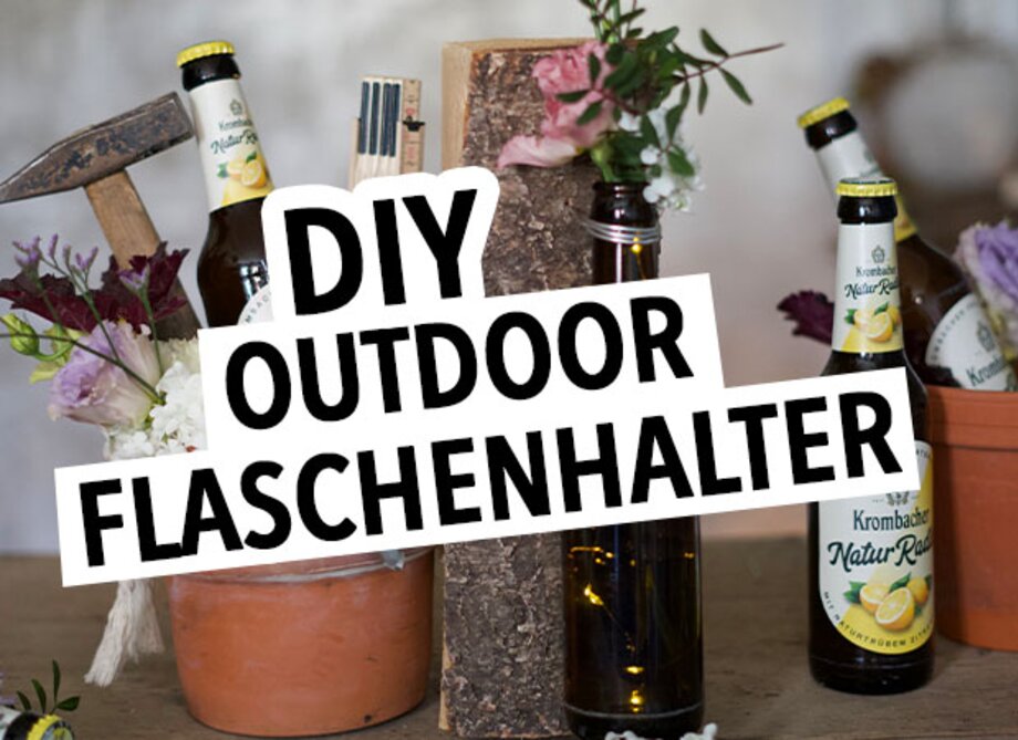 Zwei Flaschen Krombacher Natur-Radler stehen in einem Blumentopf und einem Holzblock. Ein Holzhammer und Blumen sind ebenfalls sichtbar. Der Text 'DIY OUTDOOR FLASCHENHALTER' ist prominent in Schwarz auf Weiß.